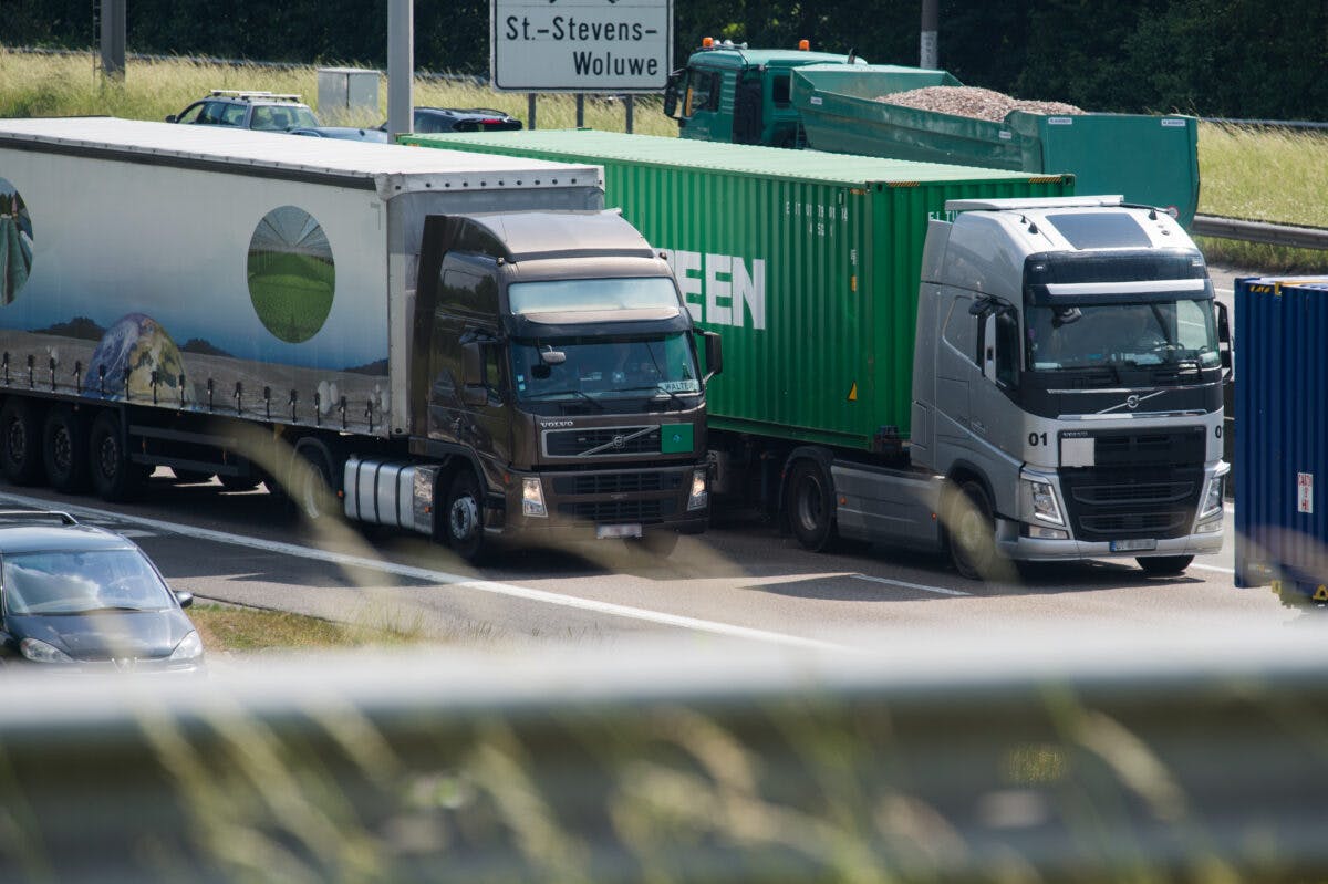 To lastebiler kjører på en motorvei med biler i forgrunnen og grønt løvverk i bakgrunnen.
