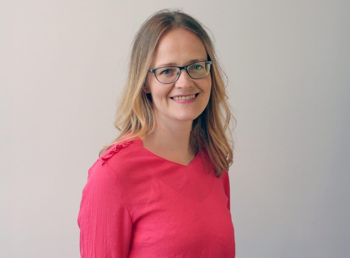 En kvinne med briller og skulderlangt lysebrunt hår, iført en rosa langermet skjorte, smiler til kameraet mot en vanlig bakgrunn.