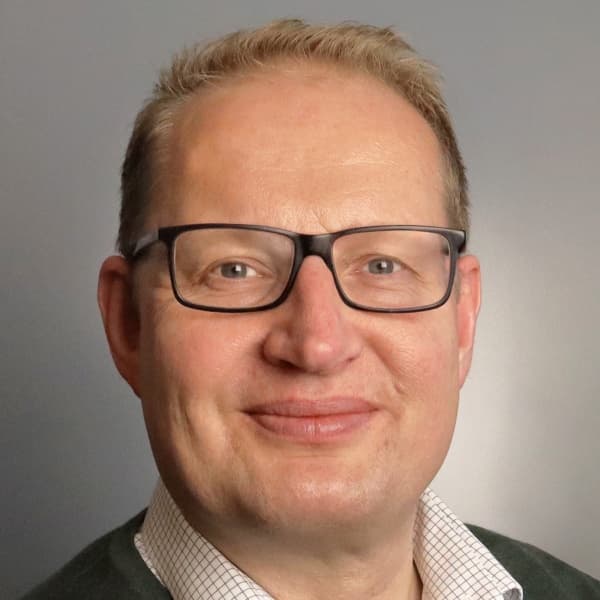 En person med briller, kort blondt hår og iført skjorte med krage smiler mot en vanlig grå bakgrunn.