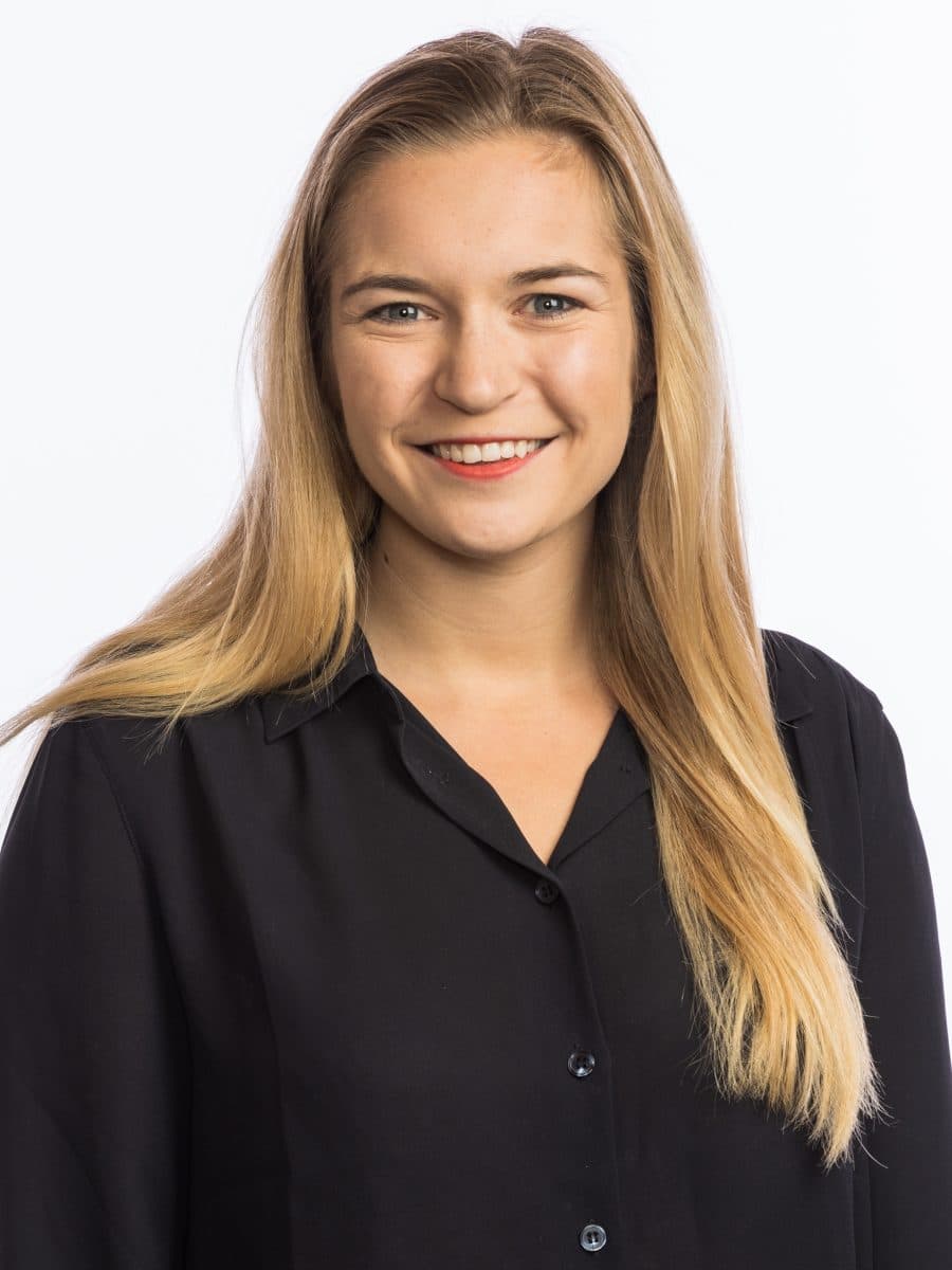 En ung kvinne med langt, blondt hår og svart skjorte smiler til kameraet mot en enkel, hvit bakgrunn.