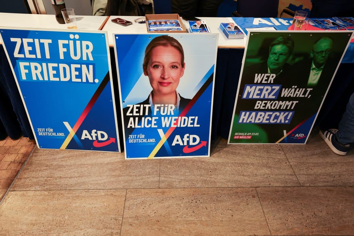 På gulvet henger tre politiske plakater fra partiet AfD i Tyskland, med slagord og et portrett av Alice Weidel.