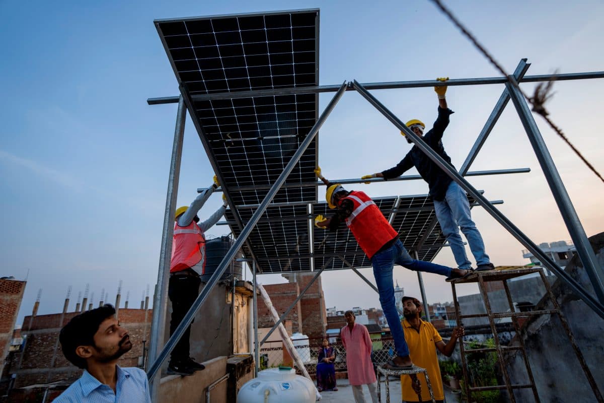 Flere arbeidere iført sikkerhetsutstyr monterer solcellepaneler på en metallkonstruksjon på toppen av en bygning, mens tilskuere står i nærheten i et urbant miljø.