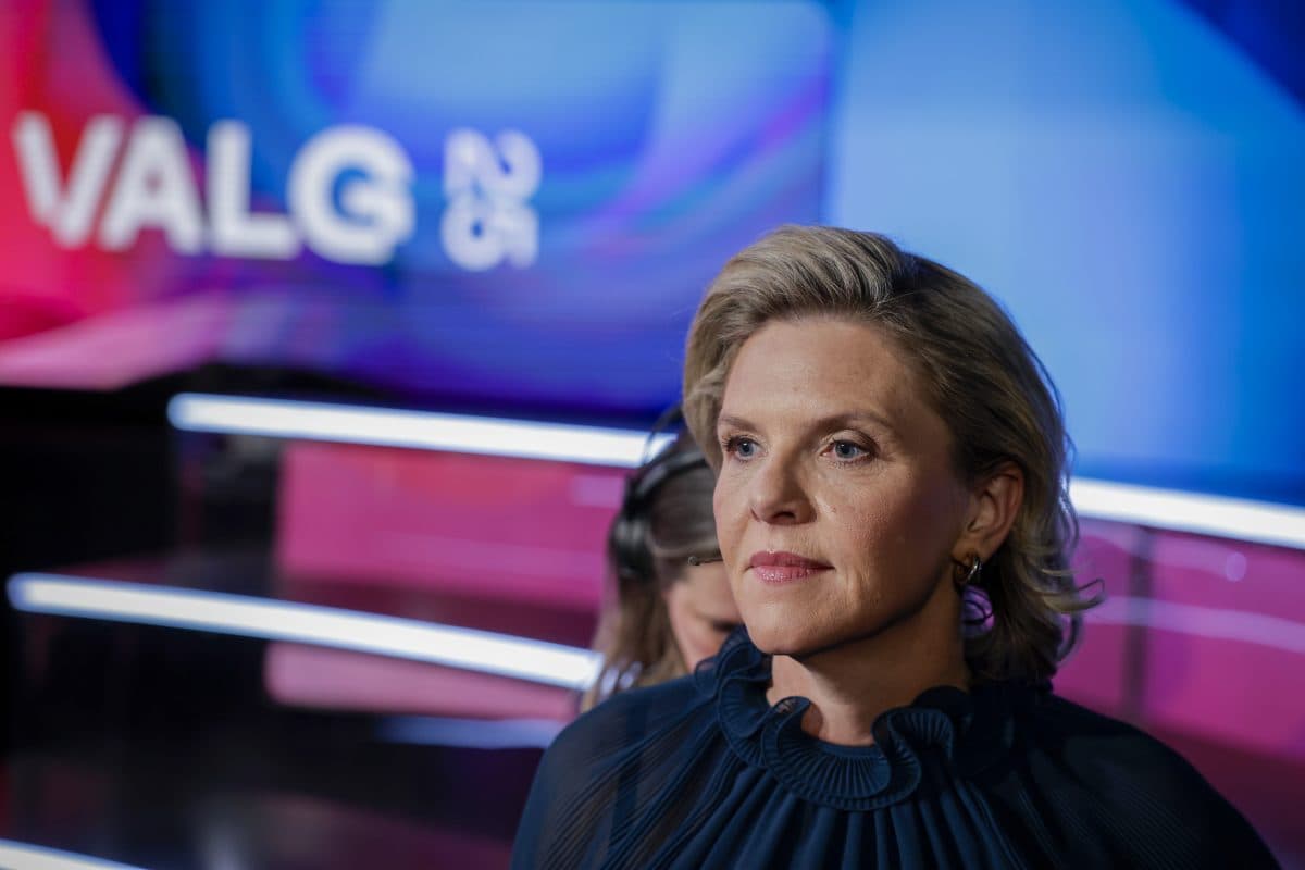 En kvinne med kort, blondt hår står foran en fargerik bakgrunn med ordet "VALG" delvis synlig. Hun har på seg en mørk, plissert topp og ser fremover med et alvorlig uttrykk.