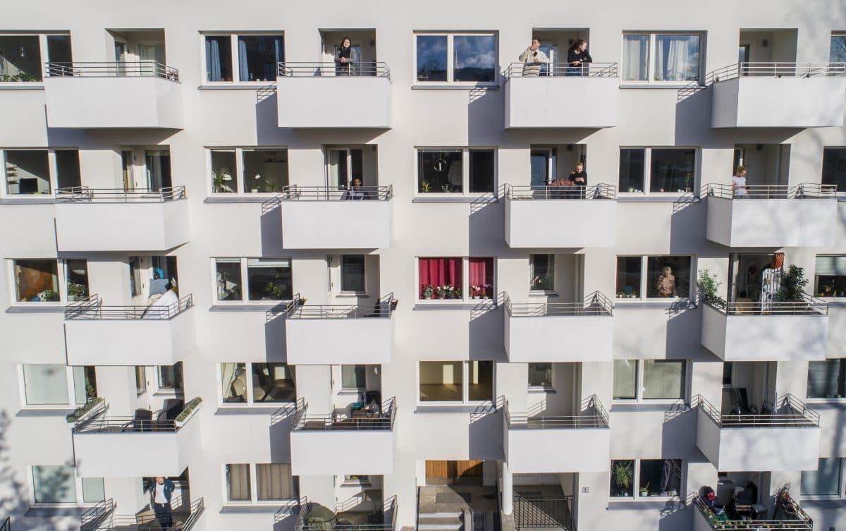 En hvit bygård med flere balkonger, hver med mennesker som står eller ser ut, og ett vindu med røde gardiner i midten.