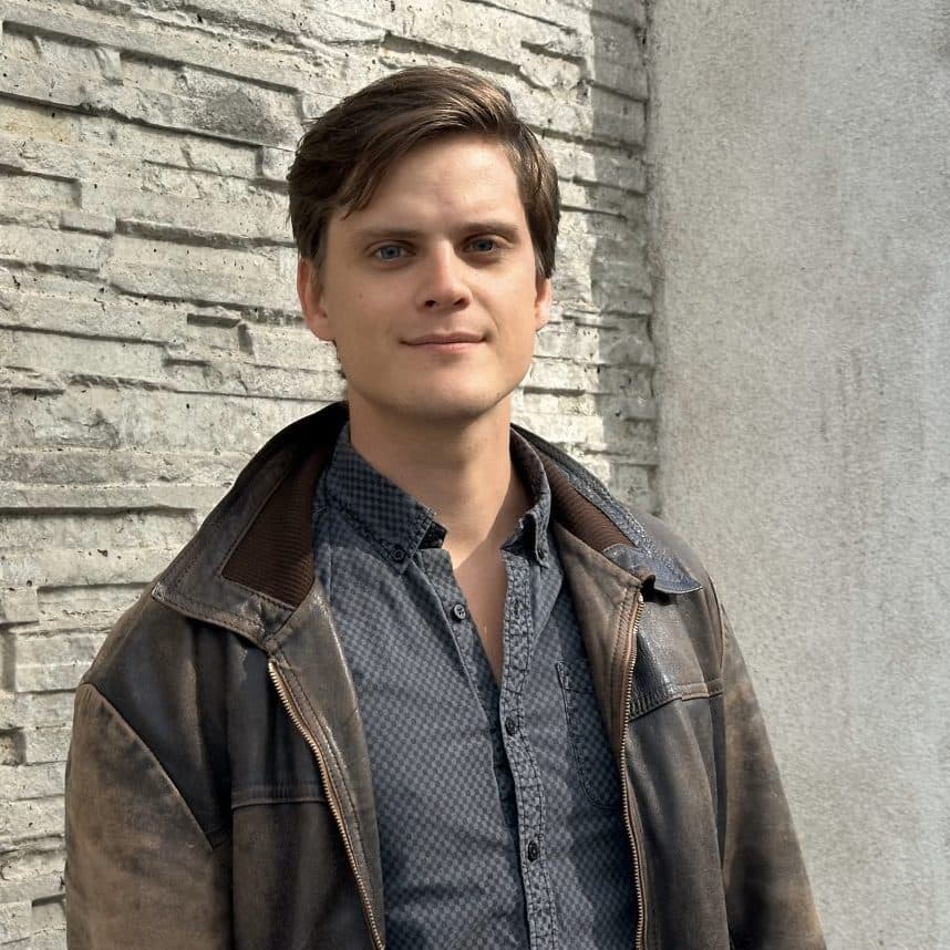 En ung mann i brun jakke og mønstret skjorte står utenfor mot en strukturert steinmur og ser inn i kameraet.