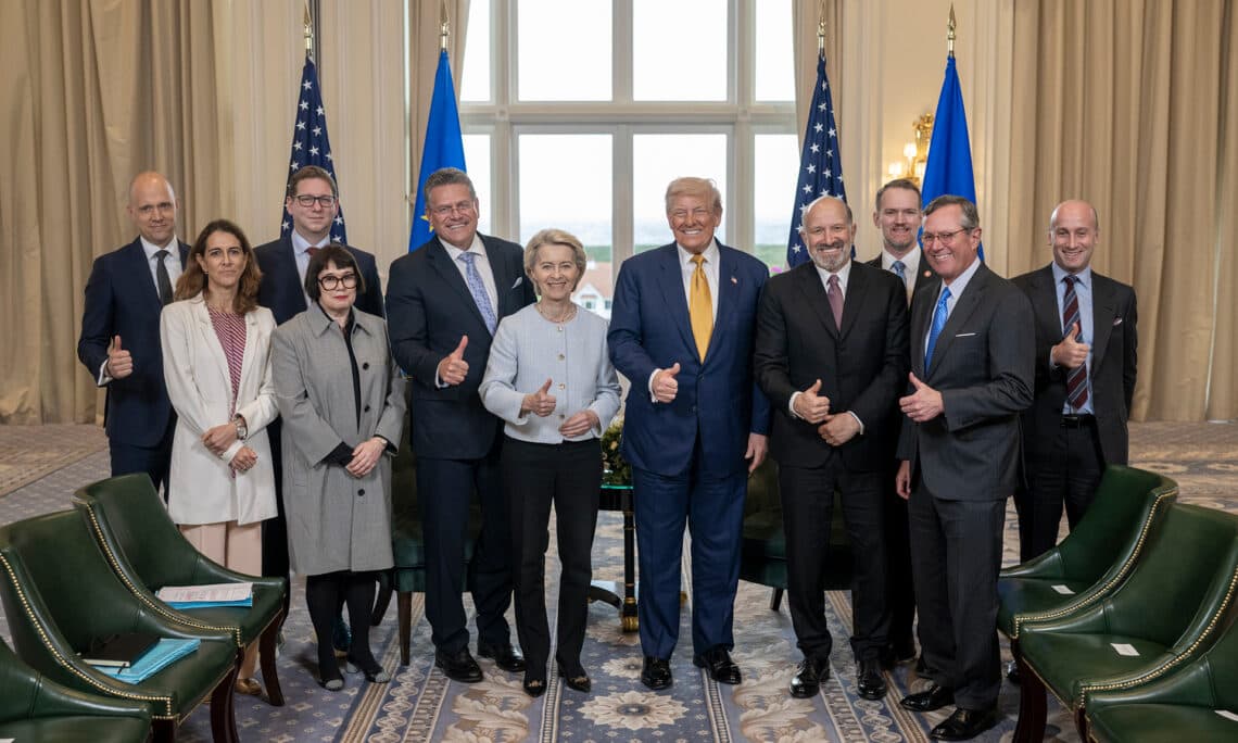 En gruppe tjenestemenn og tidligere president Donald Trump står innendørs foran store vinduer, alle smiler og gir tommelen opp, med to amerikanske og to EU-flagg i bakgrunnen.