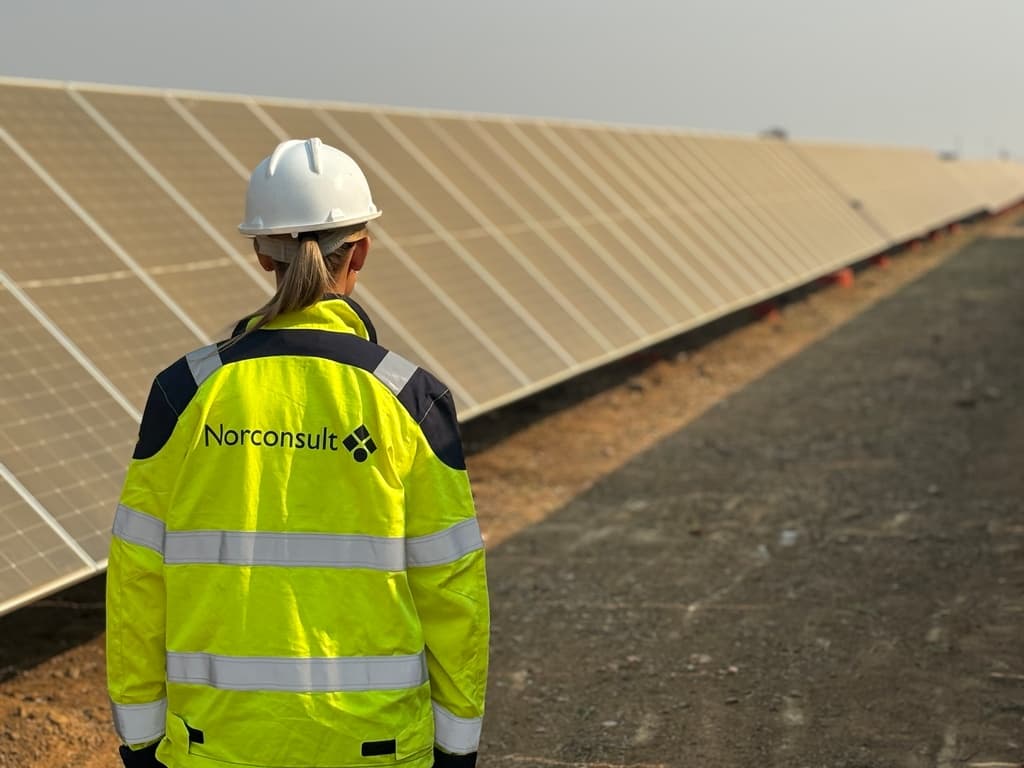 En person i varseljakke og hjelm står foran en stor oppstilling av solcellepaneler på en solrik dag.