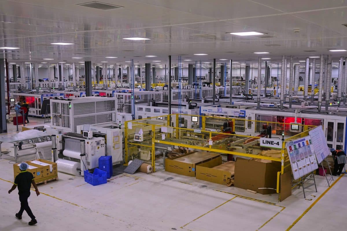 En romslig, sterkt opplyst fabrikk med rader av maskiner, arbeidsstasjoner og transportbånd. Noen få arbeidere i uniformer er synlige på det rene fabrikkgulvet.