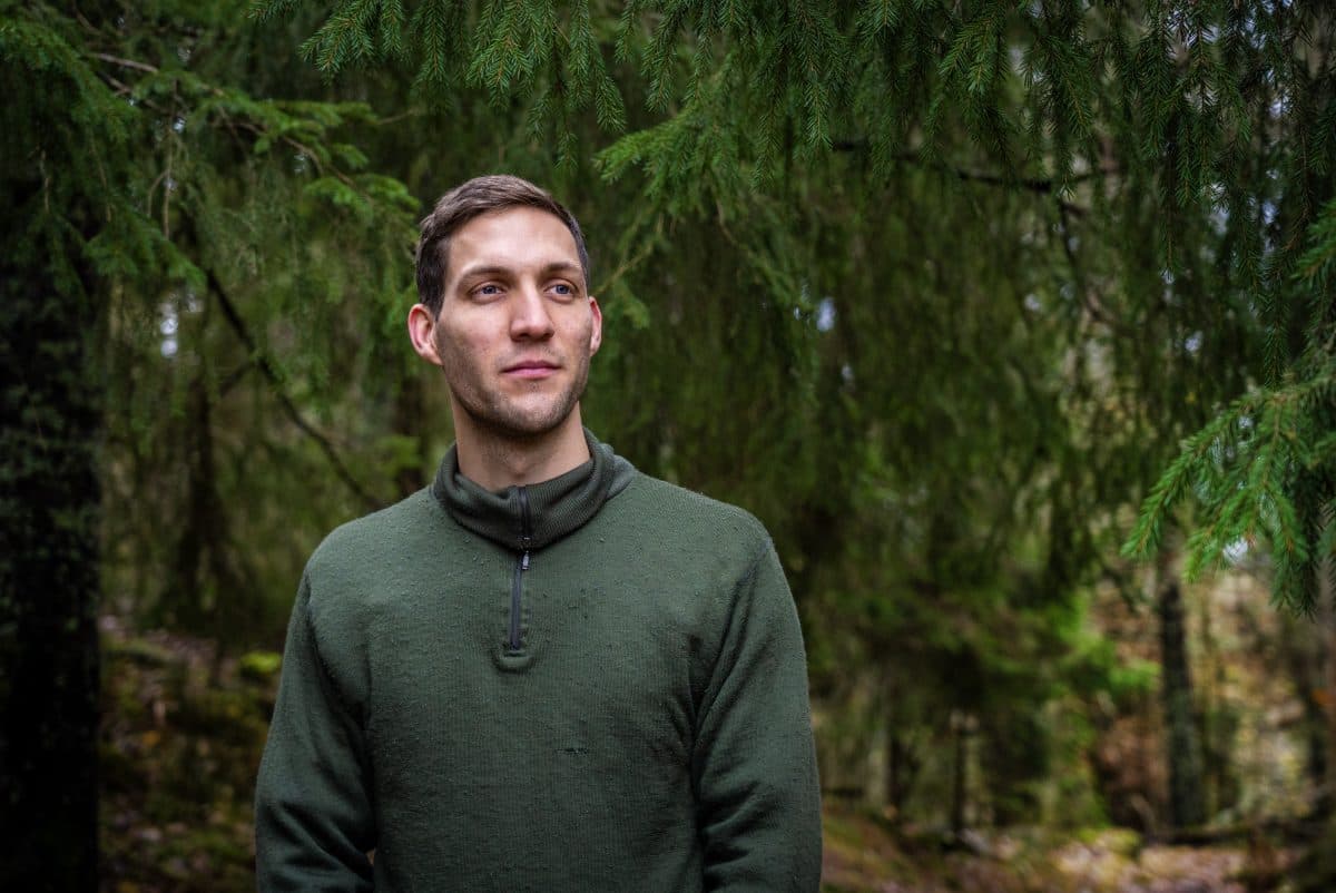 En mann i grønn genser står utendørs, omgitt av tette, grønne trær og løvverk i et skogsmiljø.