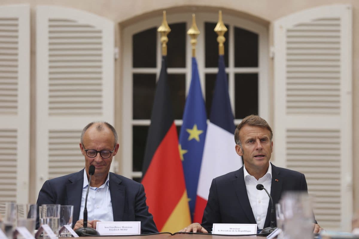 To menn sitter ved et bord foran tyske, europeiske og franske flagg og snakker på et formelt arrangement med mikrofoner og navneskilt.