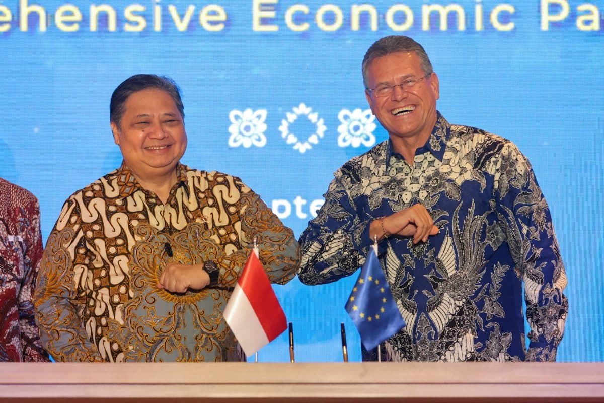 To menn i batikkskjorter hilser på hverandre med albuestøt foran indonesiske flagg og EU-flagg under et offisielt arrangement.