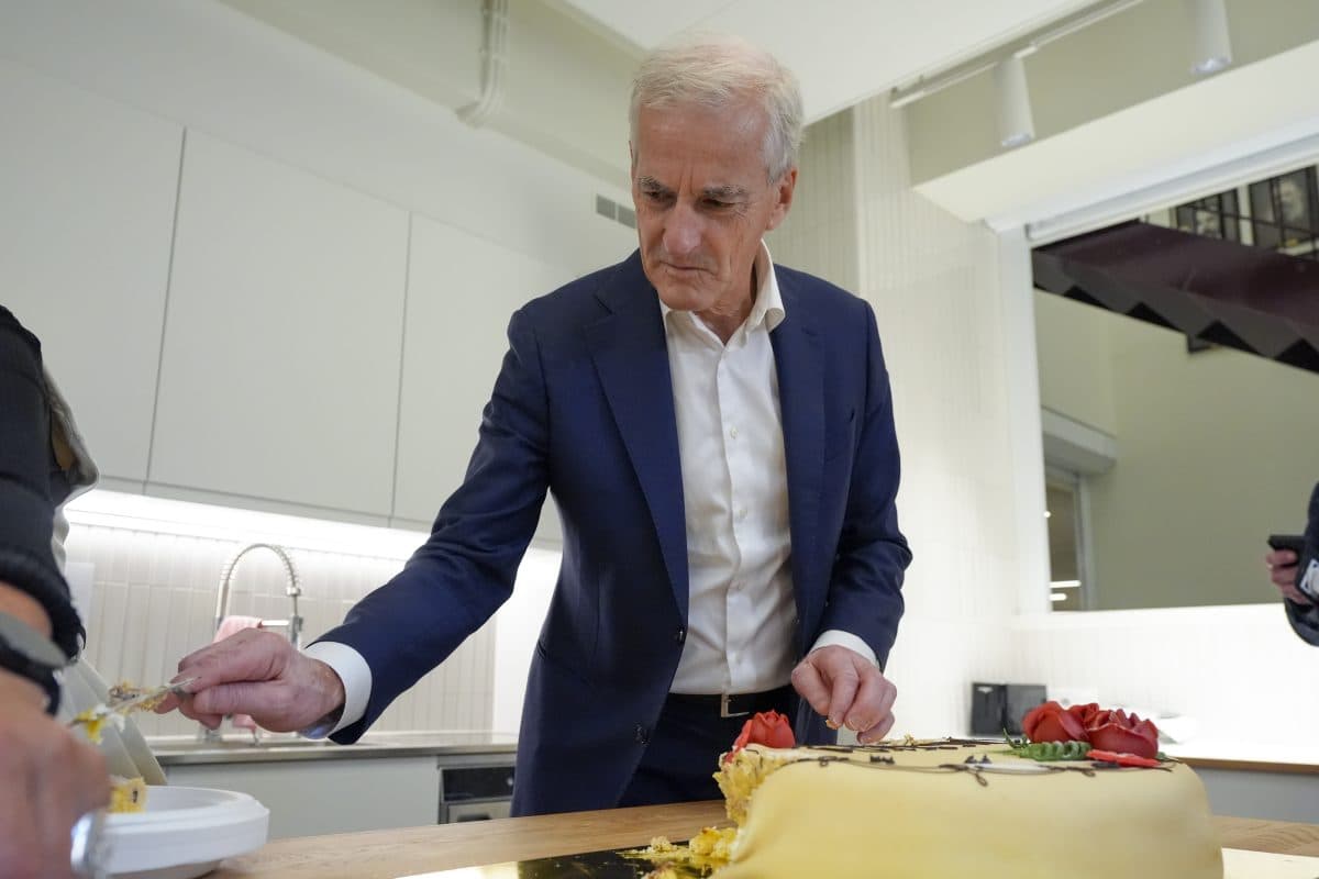 En mann i blå dress står ved et bord og strekker seg etter et stykke gul kake med røde dekorasjoner i en moderne kjøkkeninnredning.