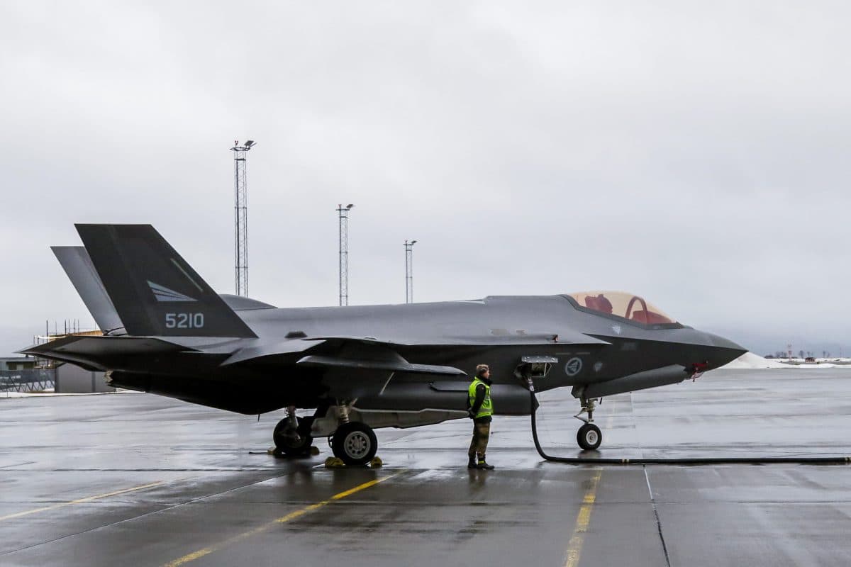 En person i refleksvest står i nærheten av et parkert F-35 jagerfly som tankes opp på en våt flyplass under en overskyet himmel.