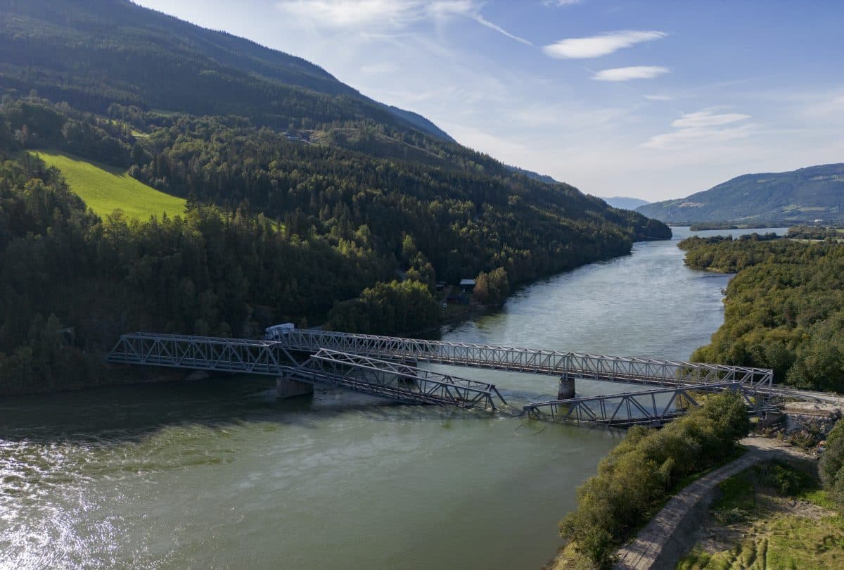 En delvis sammenrast jernbanebro av metall spenner over en bred elv, omgitt av skogkledde åser og en skyfri himmel.