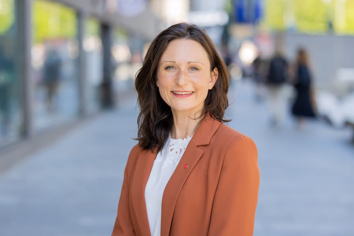 Kvinne med skulderlangt, brunt hår iført rustfarget blazer og hvit topp står utendørs i en bygate og smiler til kameraet.