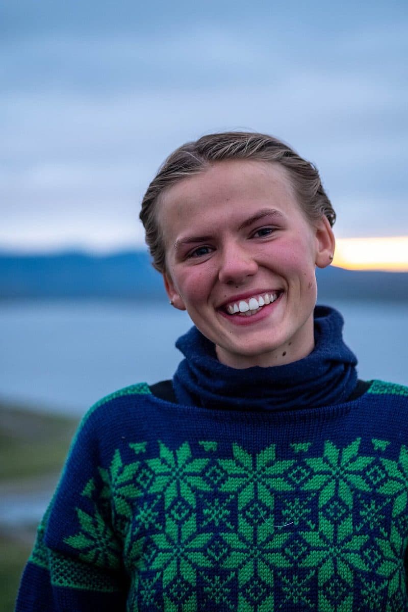 En person med kort, lysebrunt hår smiler til kameraet, iført en blå og grønnmønstret genser, utendørs i nærheten av vann med åser og en overskyet himmel i bakgrunnen.