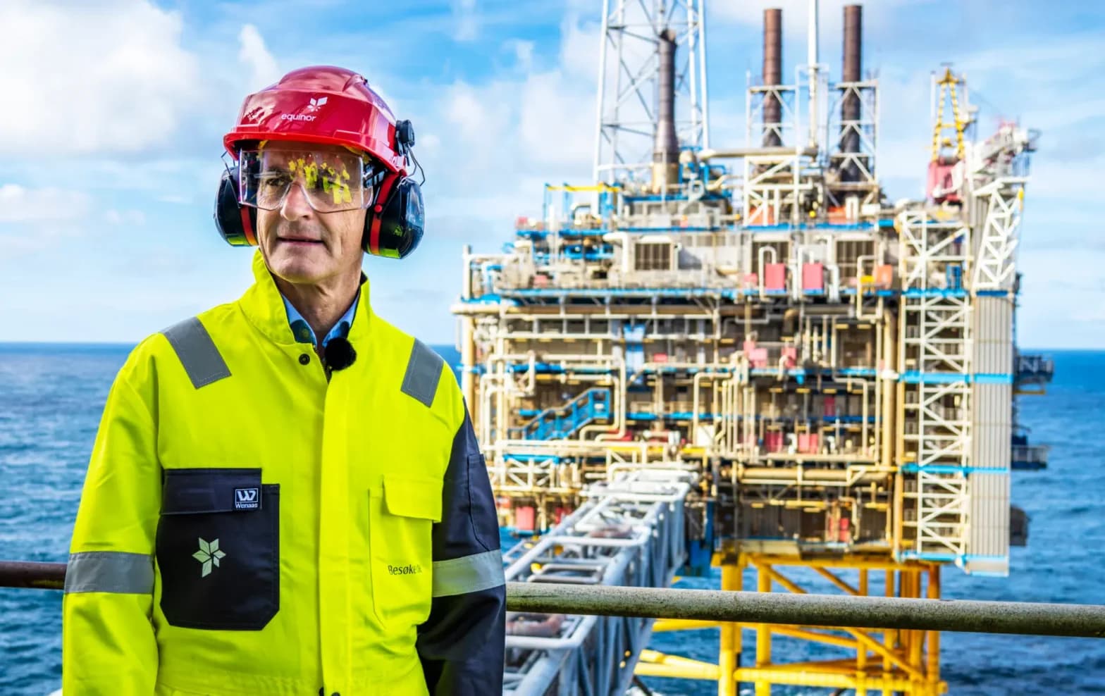 En arbeider iført sikkerhetsutstyr står på en oljeplattform til havs, med industristrukturer og havet synlig i bakgrunnen.