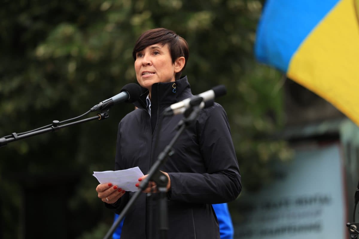 En kvinne snakker i en mikrofon utendørs med papirer i hånden, mens et ukrainsk flagg er synlig i forgrunnen.