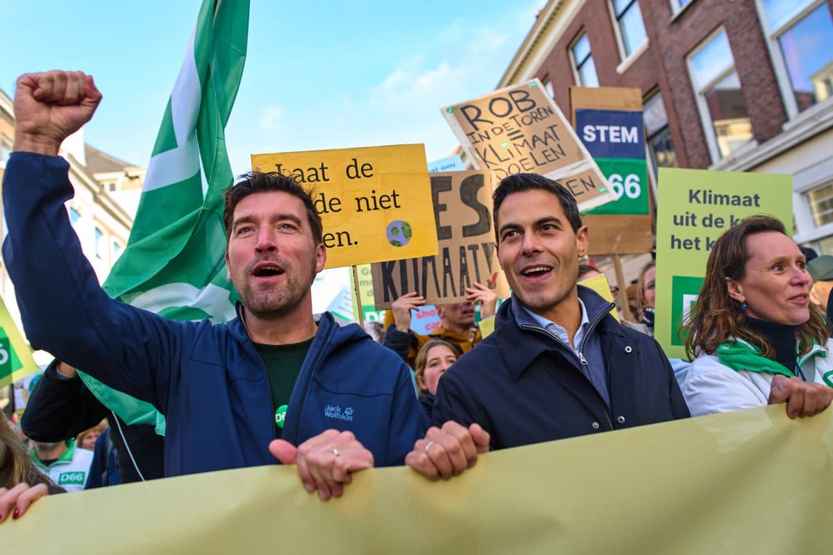 En gruppe mennesker med bannere og skilt under en klimaprotest, med to menn i front, hvorav den ene hever knyttneven, i en bygate.