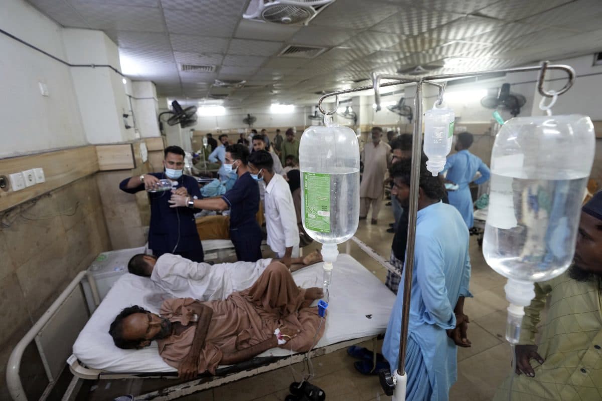 Et overfylt sykehusrom med flere pasienter som ligger i senger og får drypp, med medisinsk personale rundt seg og mange mennesker rundt seg.