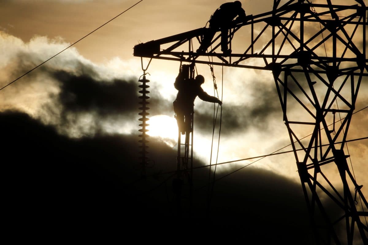 To arbeidere utfører vedlikehold på en høyspentmast i solnedgang, med skyer og sol i bakgrunnen.