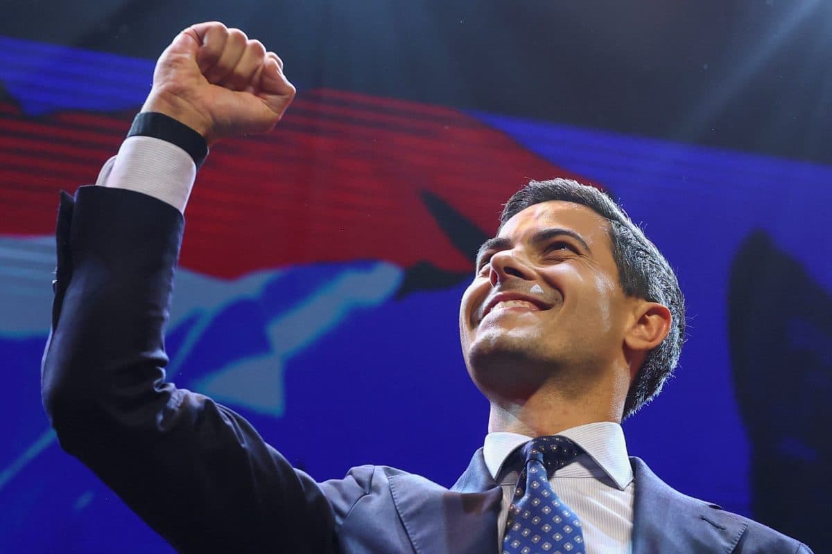 En mann i dress hever neven og smiler på scenen, med en blå og rød bakgrunn bak seg.