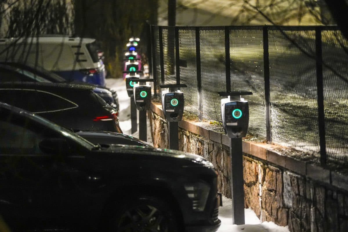 Flere elbiler står parkert på rekke og rad om natten, koblet til ladestasjoner langs et snødekt fortau ved siden av et gjerde.