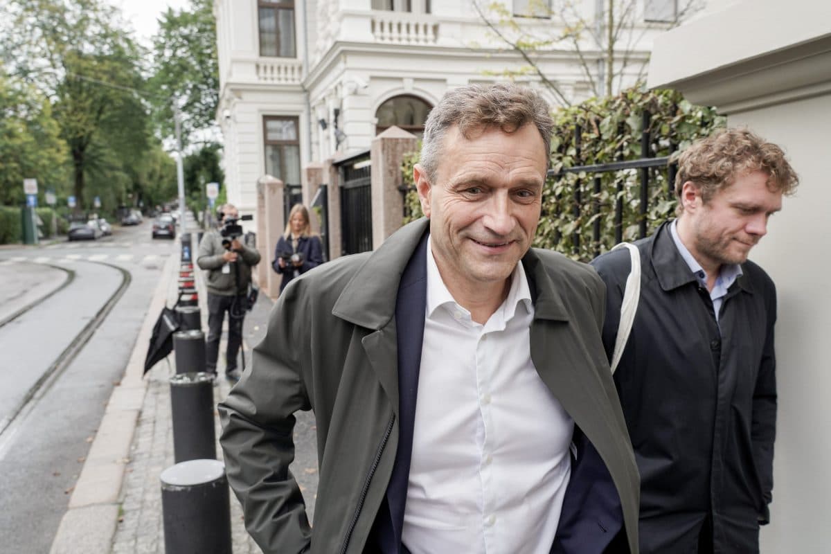 En mann i hvit skjorte og mørk jakke går på et fortau i en by, sammen med andre, med trær og bygninger i bakgrunnen.