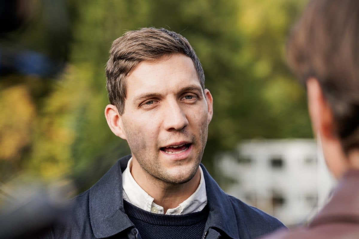 En mann med kort, brunt hår og mørk jakke snakker utendørs med en annen person; i bakgrunnen er det uskarpt grønt.