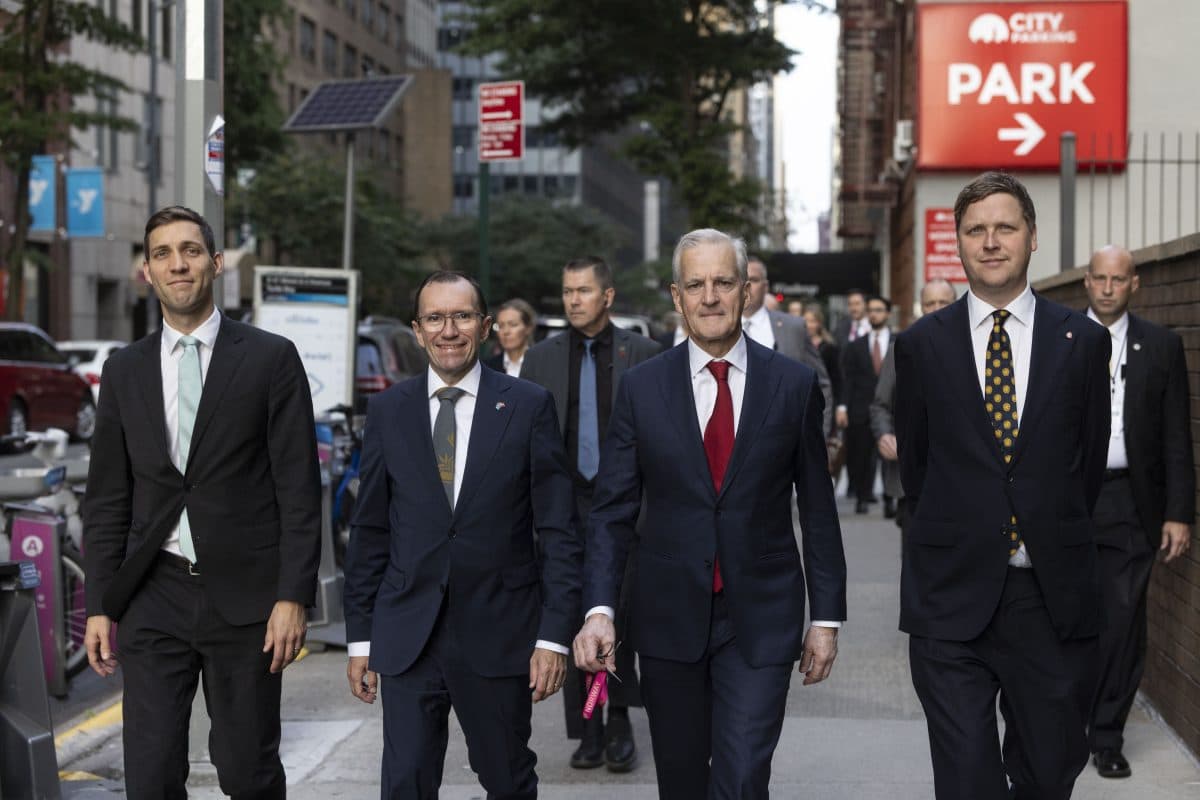 En gruppe menn i dress går sammen på et fortau i en by, med bygninger, parkerte sykler og et parkeringsskilt synlig i bakgrunnen.