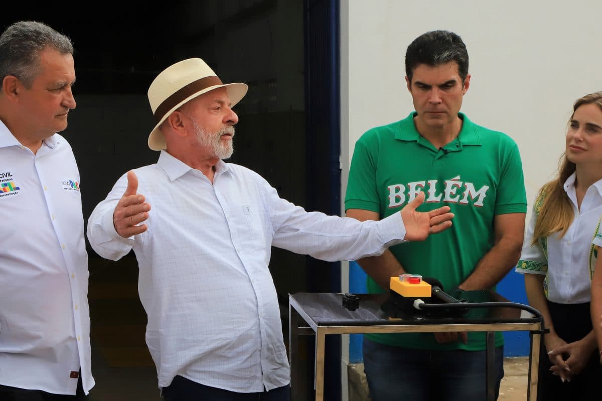 En mann i hvit skjorte og hatt gestikulerer mens han snakker til tre personer ved et lite bord med en gul og en rød knapp.