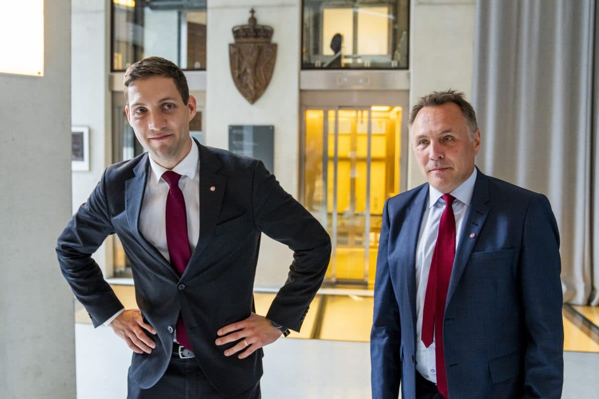 To menn i dress og rødt slips står innendørs i nærheten av en heis og en vegg med et våpenskjold.