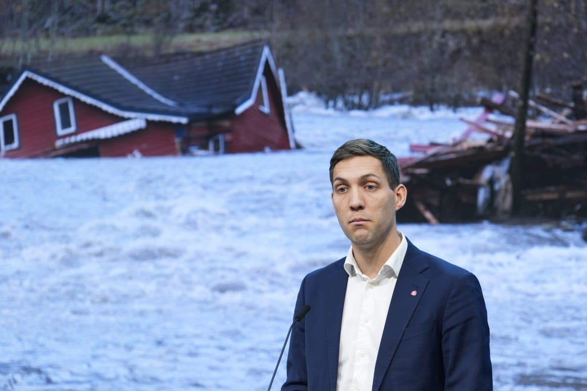 En mann i dress står på et podium foran et bilde som viser et oversvømt område med ødelagte hus.