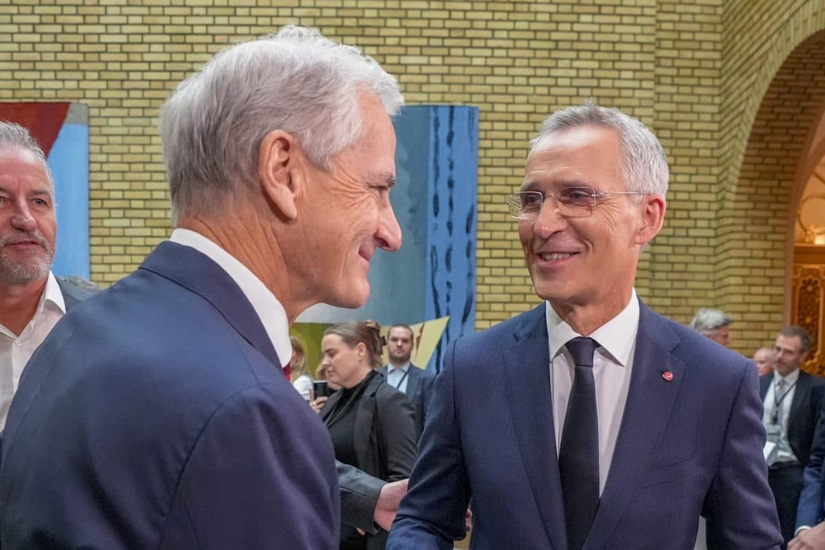 To menn i dress hilser på hverandre og smiler i en formell innendørssetting, med flere mennesker og abstrakt kunst synlig i bakgrunnen.