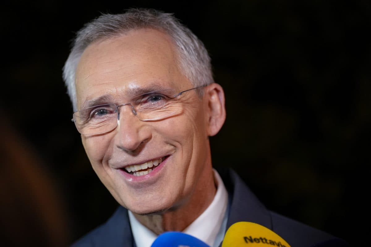 En eldre mann med briller og grått hår smiler mens han snakker til pressen, med flere fargede mikrofoner synlige foran seg.