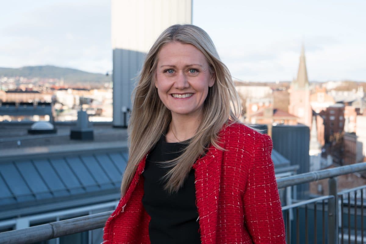 En kvinne med langt blondt hår, iført rød jakke og svart topp, står på et hustak med et bylandskap i bakgrunnen.