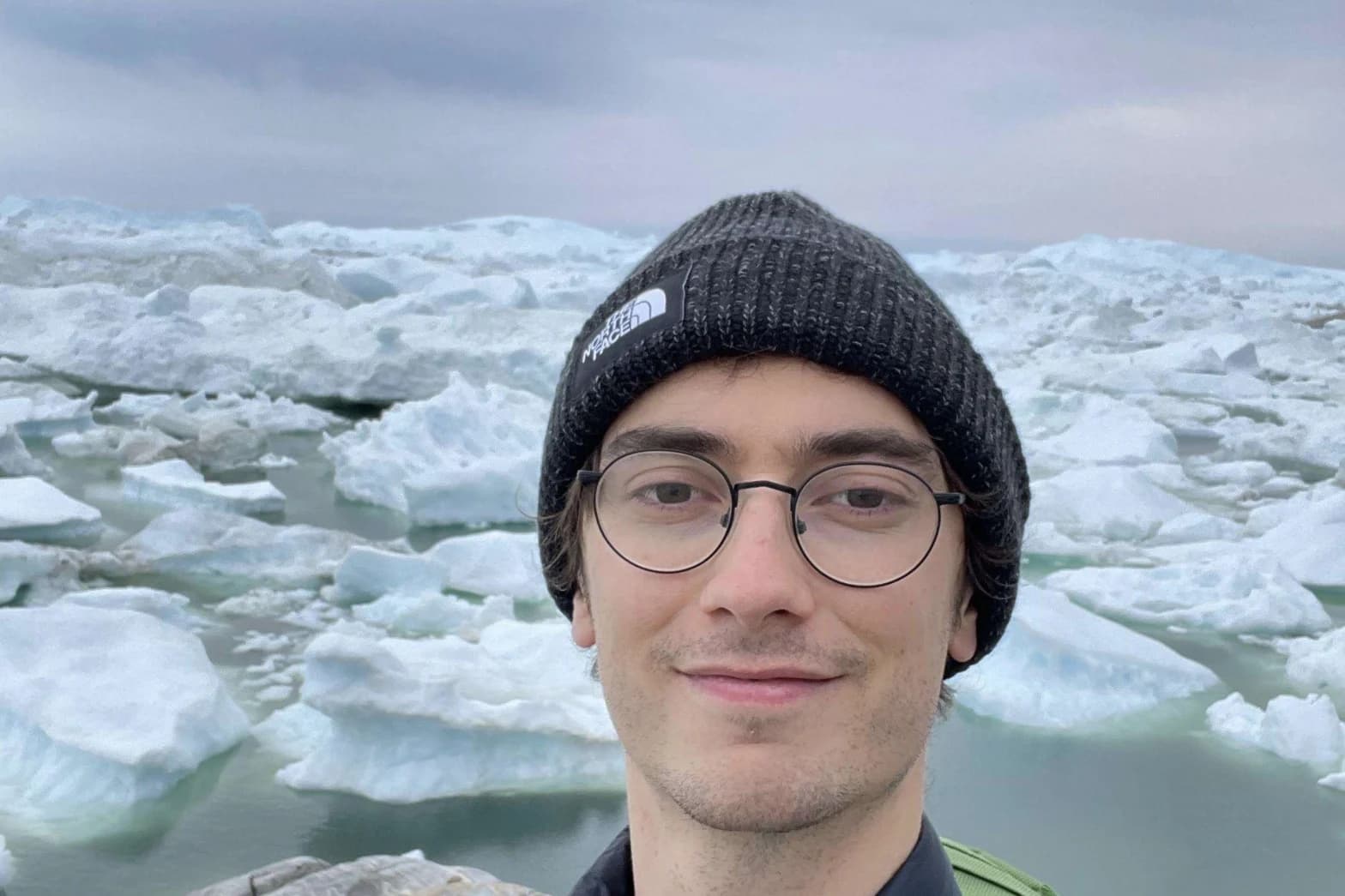En person med briller og svart strikkelue står foran et islandskap med flytende isfjell og overskyet himmel.