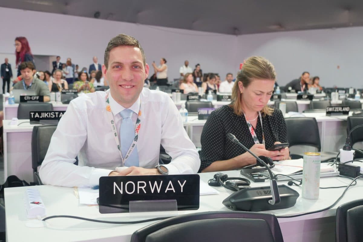 En mann sitter smilende bak et "Norway"-skilt på en internasjonal konferanse, mens en kvinne ved siden av ham ser på telefonen sin. Andre delegater sitter i bakgrunnen.