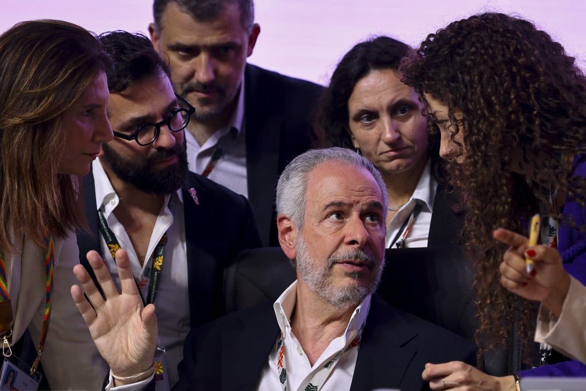 En sittende mann med grått hår gestikulerer mens fem kolleger står rundt ham, alle engasjert i en alvorlig diskusjon.