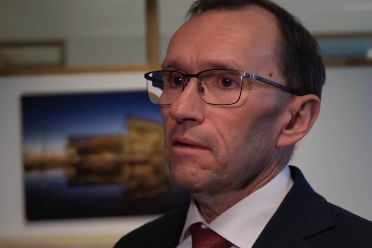 En mann i dress og briller står innendørs, med et uskarpt fotografi av en bygning og vann i bakgrunnen.