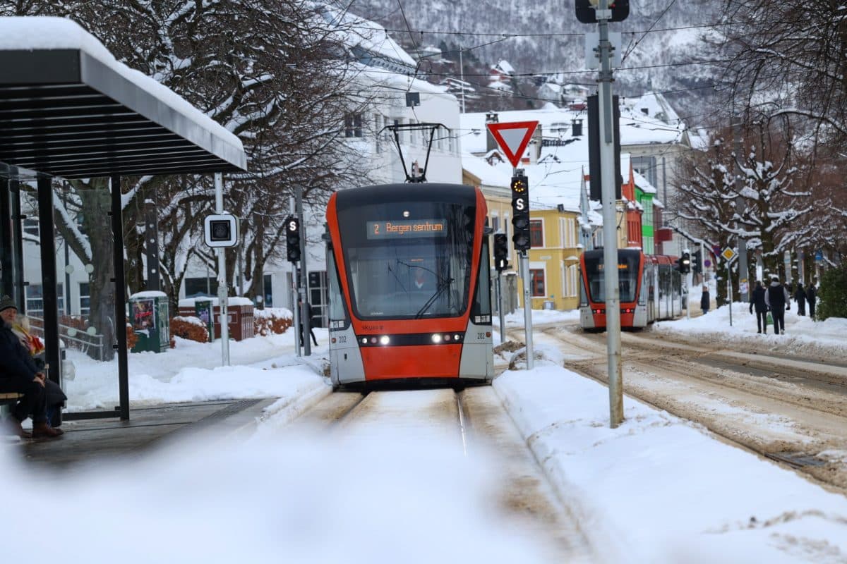 En trikk nærmer seg en snødekt stasjon i en by, med en annen trikk synlig i bakgrunnen og bygninger langs gaten.