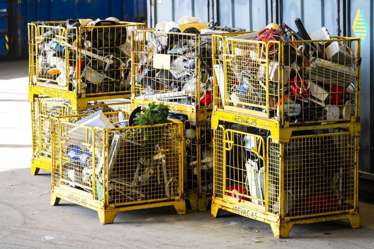 Seks gule metallbur fylt med ulike typer elektronisk avfall og skrapmaterialer er stablet sammen i et innendørs industrimiljø.