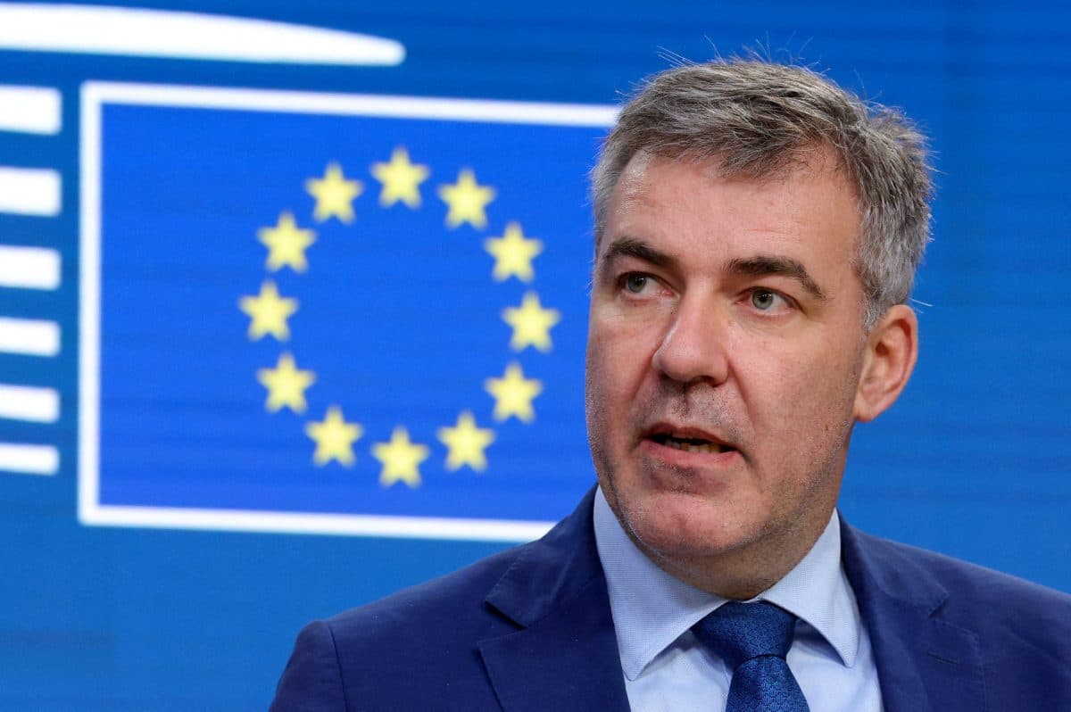 En mann i dress snakker foran en blå bakgrunn med EU-flagget med gule stjerner i en sirkel.