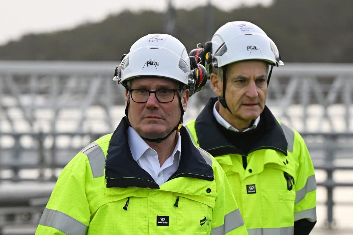 To menn iført hvite hjelmer og gule refleksvester står utendørs i nærheten av en industri- eller byggeplass.