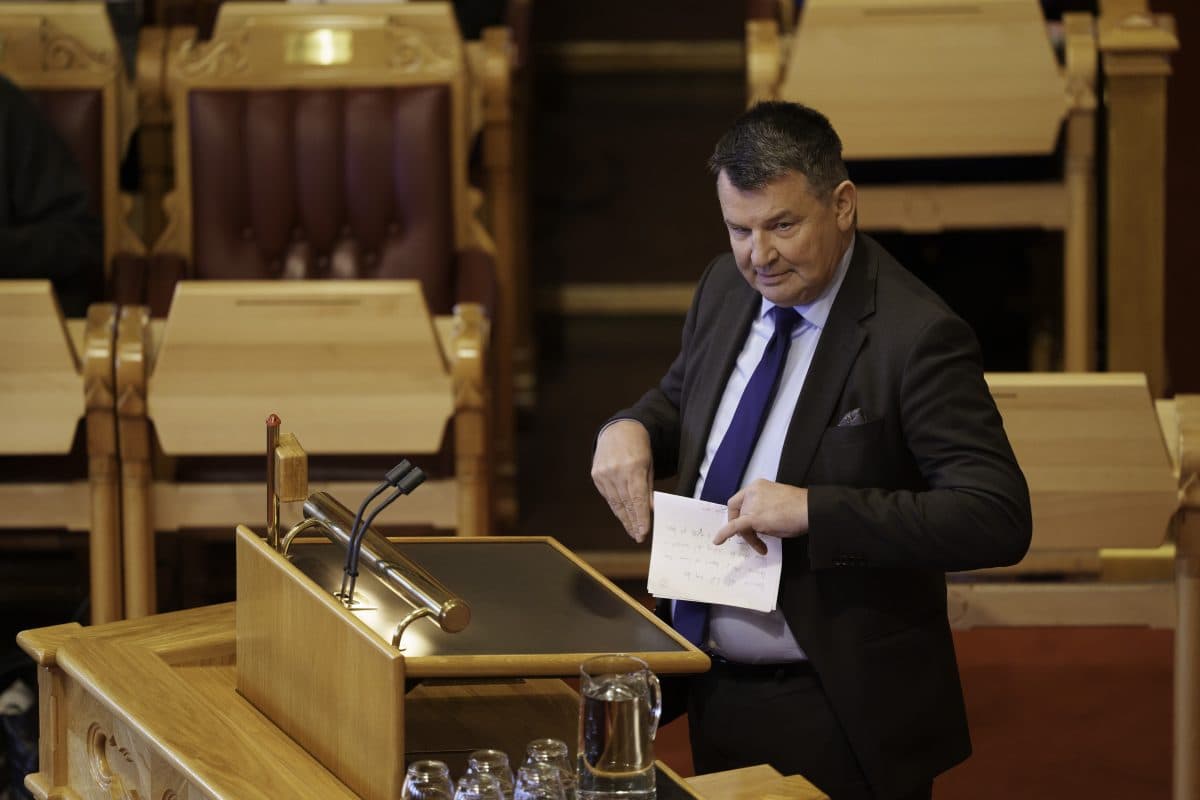 En mann i dress står på et trepodium i et stort møterom, holder et stykke papir og gestikulerer med hånden.