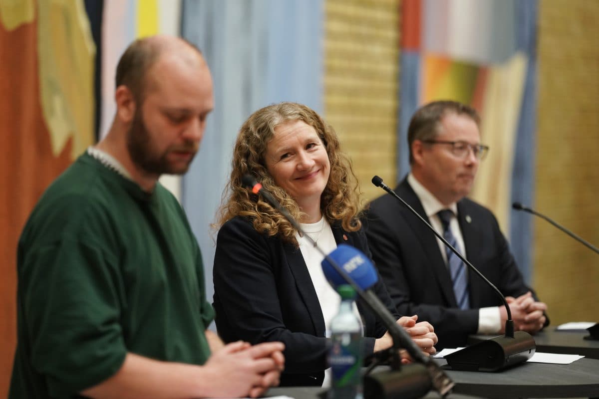 Tre personer sitter ved et bord med mikrofoner under en pressekonferanse; kvinnen i midten smiler, mens de to mennene på hver side virker fokuserte.