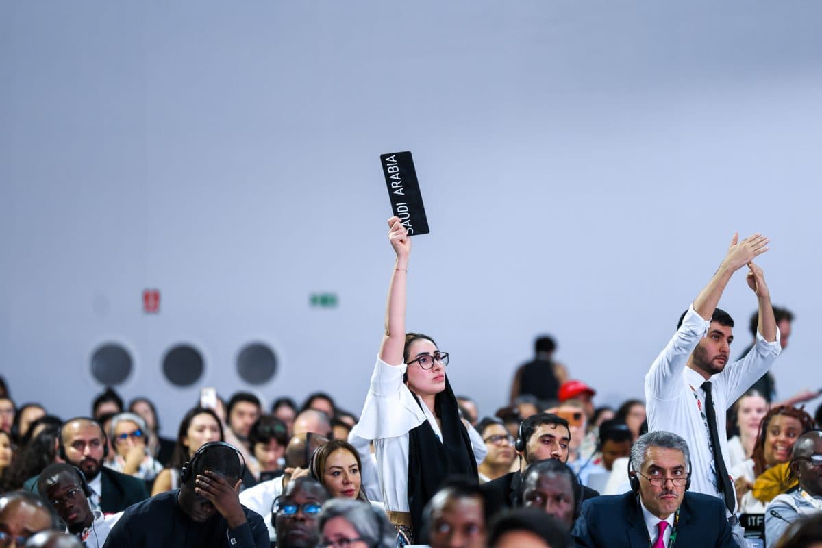 En kvinne holder opp et skilt med teksten "Saudi-Arabia" mens hun sitter blant en stor gruppe mennesker på et formelt arrangement eller en konferanse.