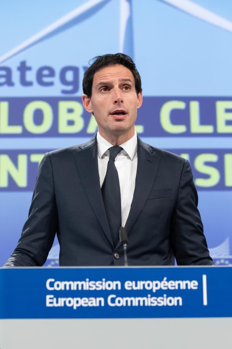 En mann i mørk dress og slips taler på et podium merket "Europakommisjonen" mot en blå bakgrunn med delvis synlig tekst.