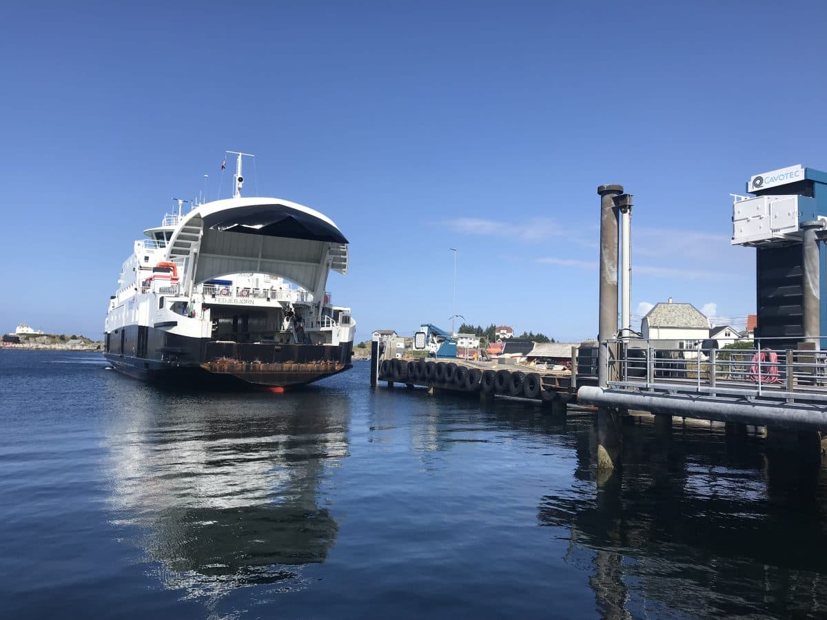 En bilferge med åpen bogrampe nærmer seg en kai en solskinnsdag, med bygninger og en klar blå himmel i bakgrunnen.