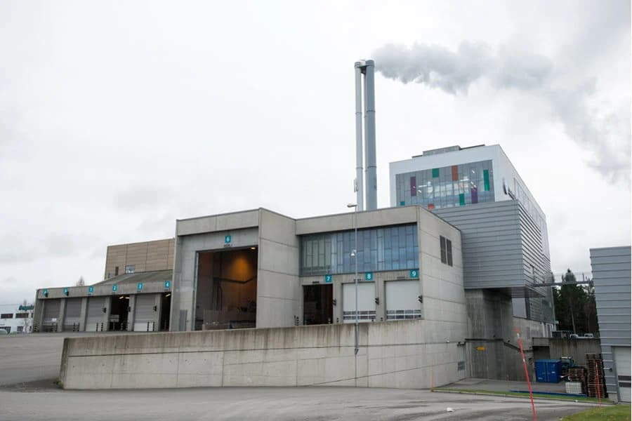 En moderne industribygning med flere lasteramper og to høye skorsteiner som sender ut hvit røyk under en overskyet himmel.